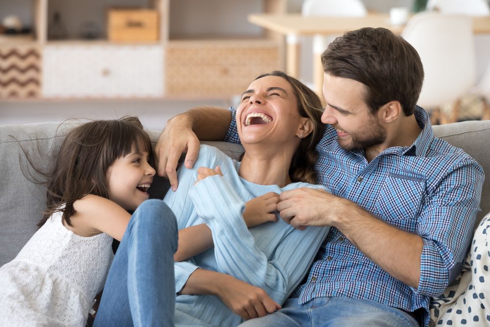 A family of three laughs together on a gray couch in a bright, cozy living room.
