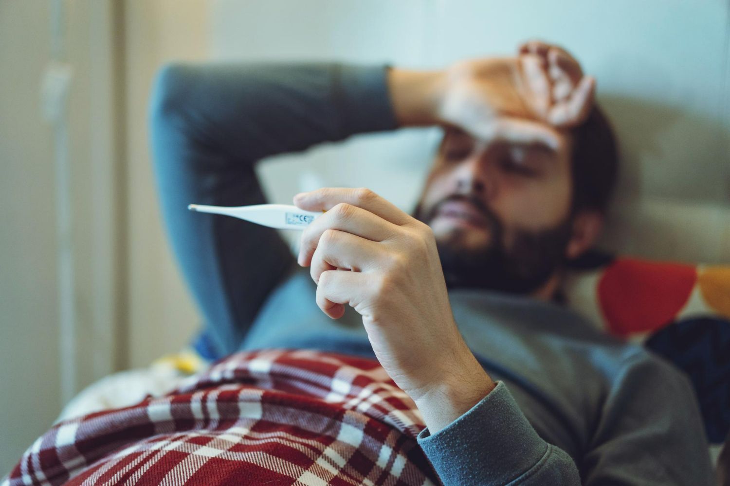 A person lying in bed, holding a thermometer and resting a hand on their forehead, appearing unwell.