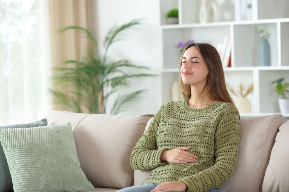 A person in a green knit sweater sits on a couch with eyes closed and hand on stomach, practicing mindfulness.