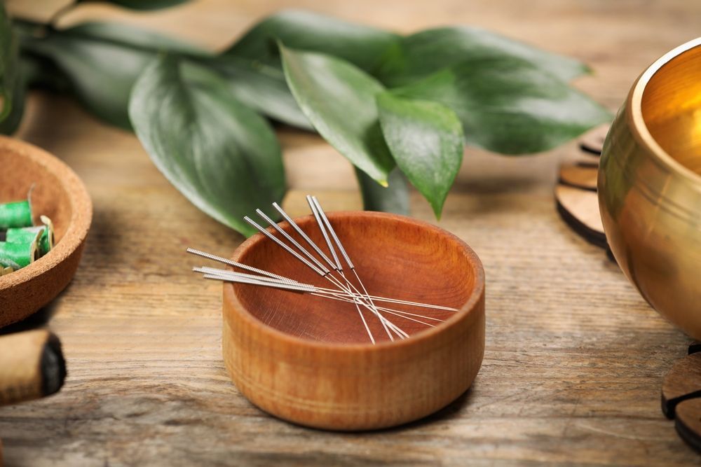 A small wooden bowl containing acupuncture needles rests on a rustic table with green leaves and a singing bowl nearby.