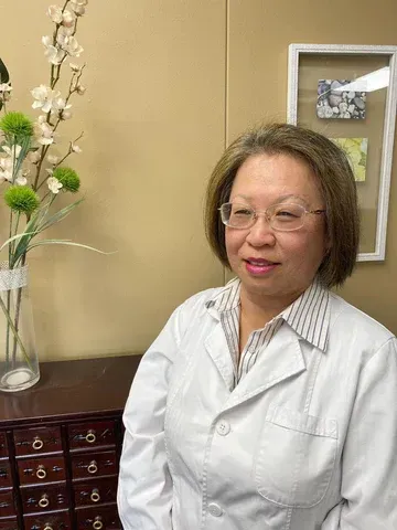 a woman in a white lab coat and glasses is standing in front of a vase of flowers .