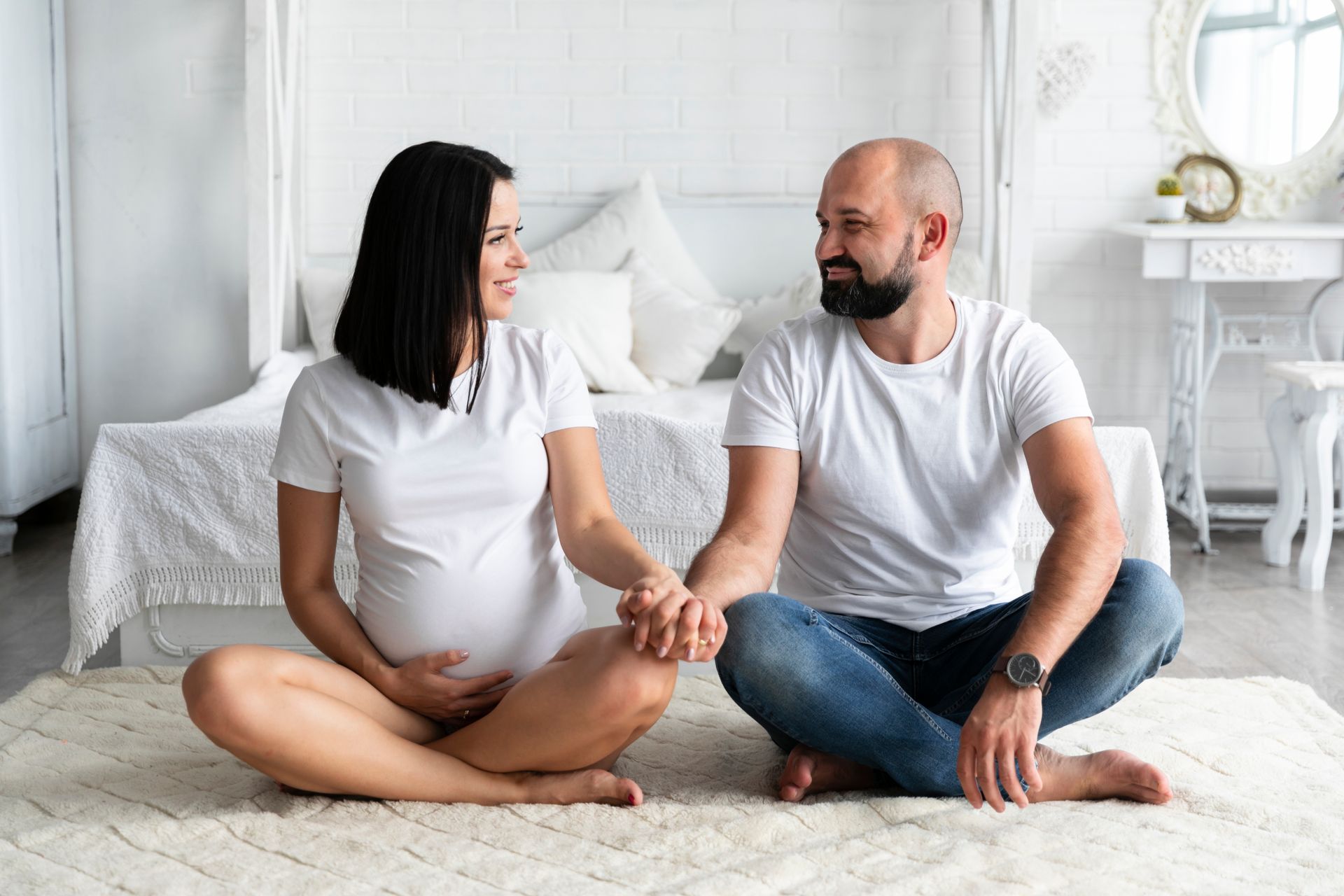 Pregnant woman and partner holding hands, sitting on a rug in a bright bedroom, looking at each other.