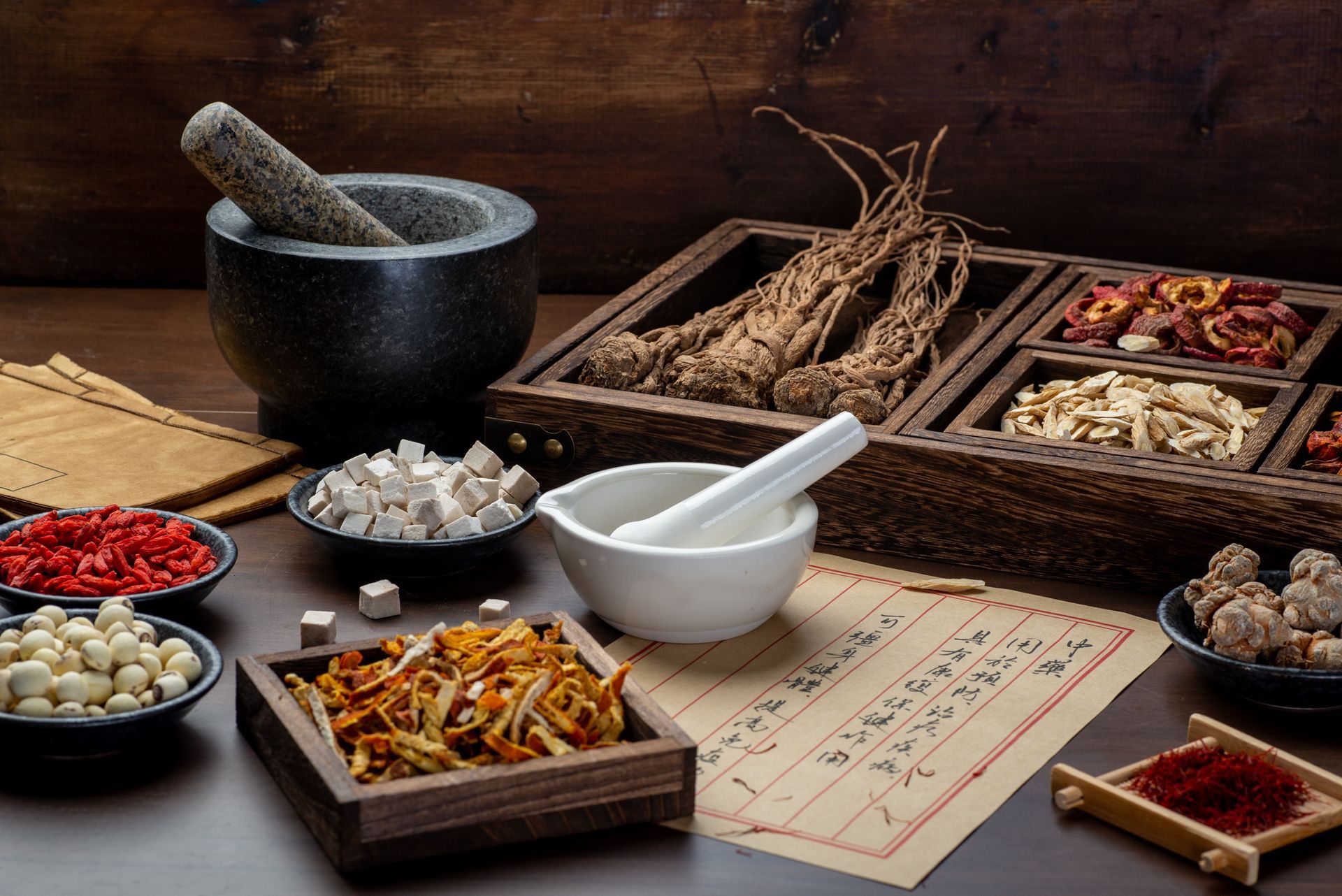 Herbs and tools for traditional medicine; mortar and pestle, wooden trays with dried roots and ingredients, script.