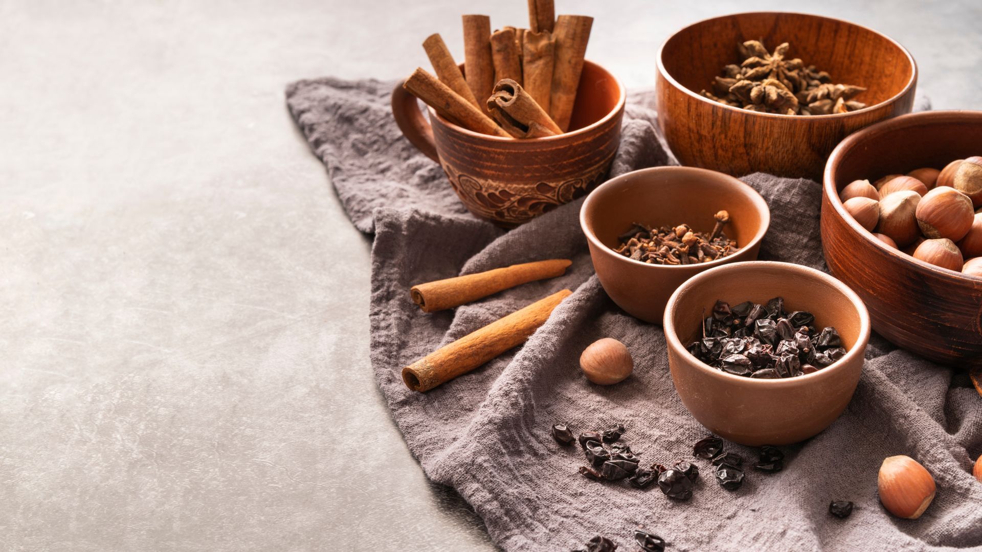 Wooden bowls filled with cinnamon sticks, cloves, star anise, nutmeg, and nuts on a gray cloth.