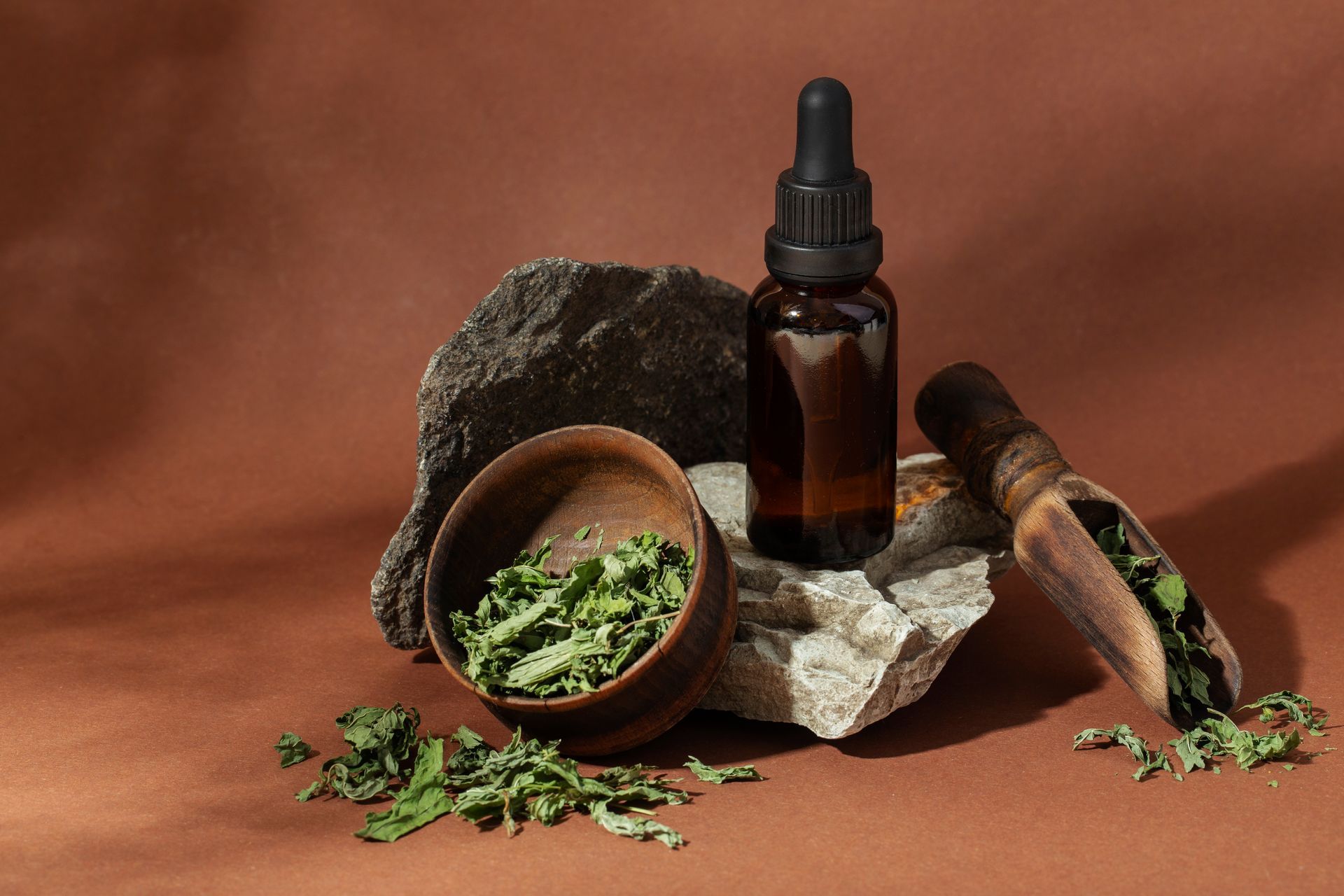 Brown dropper bottle on rock with dried herbs in a bowl and scoop against a brown backdrop.