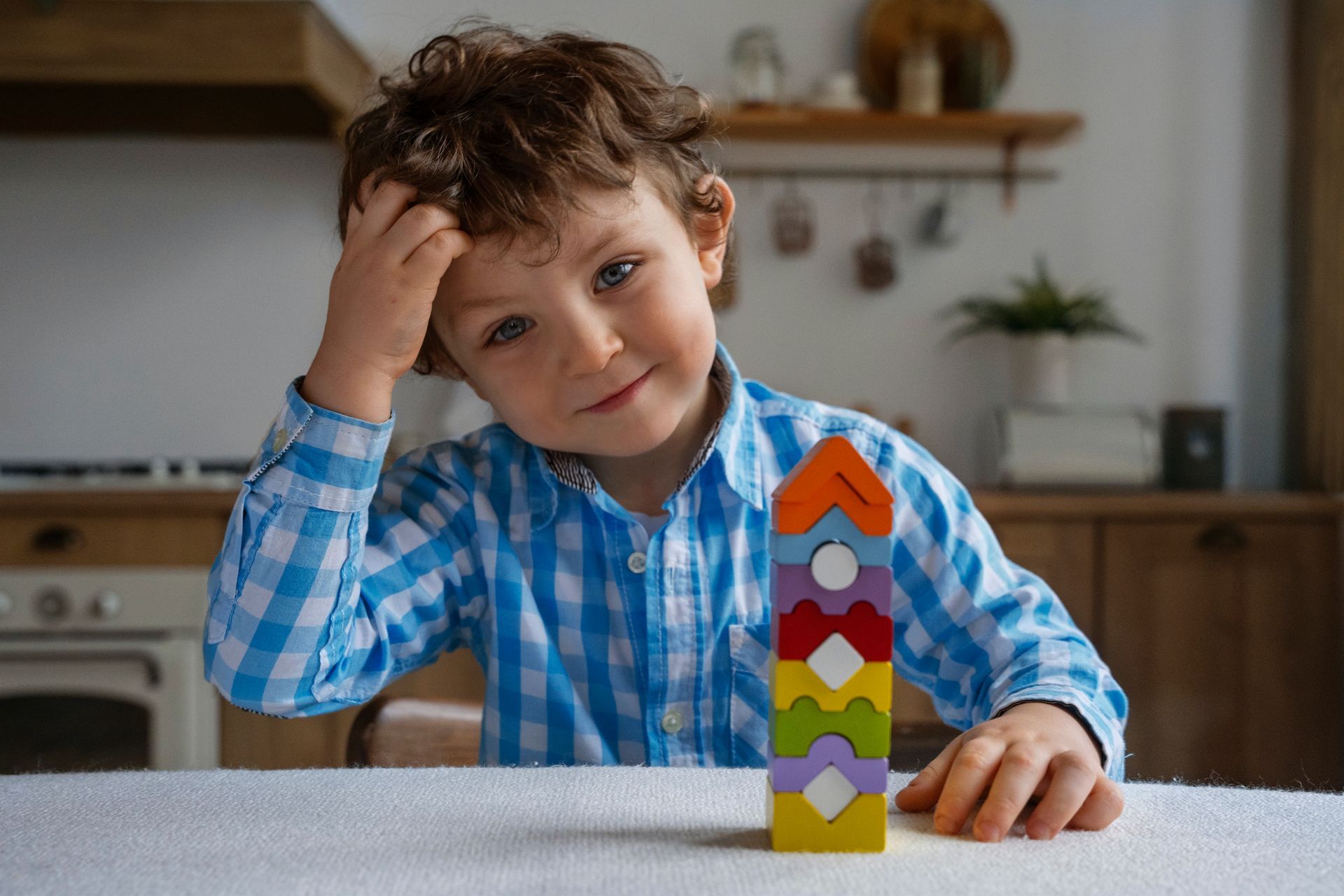 Boy with curly hair pondering a colorful wooden block tower at a table indoors.