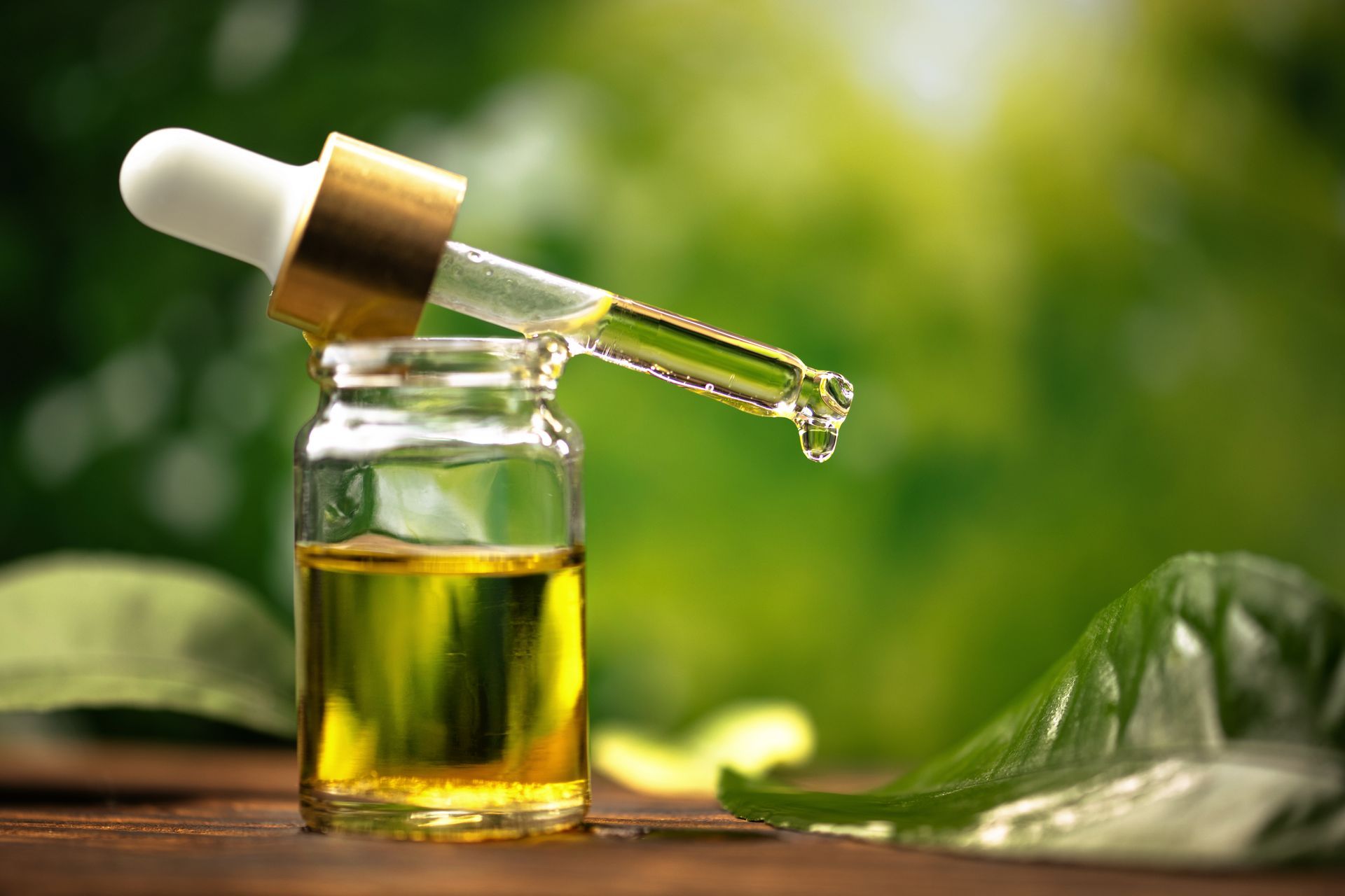 Dropper dispensing yellow liquid into a small glass bottle, on a wooden surface with green foliage background.