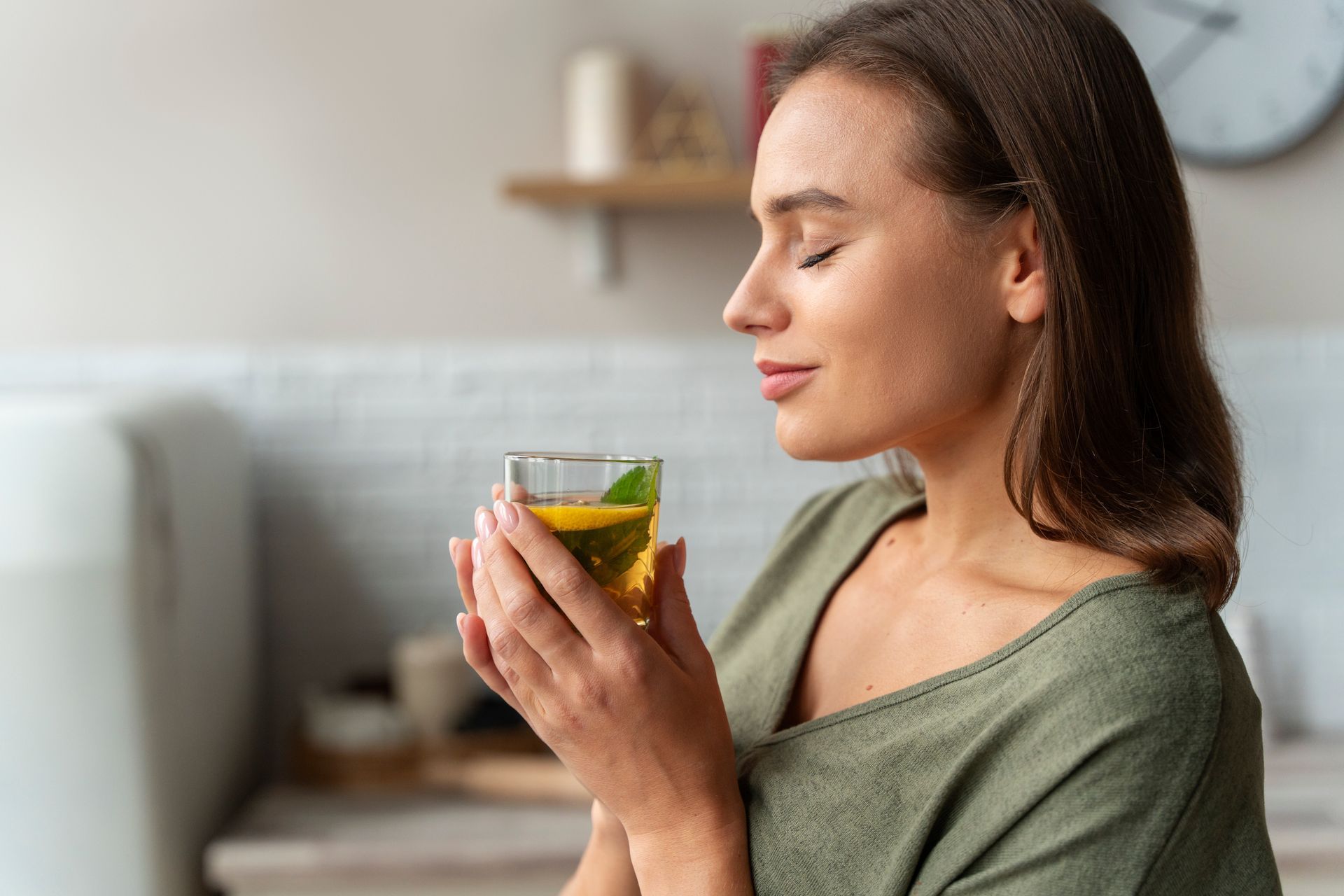 Woman in green shirt holds glass of tea, eyes closed, enjoying the aroma in a kitchen setting.