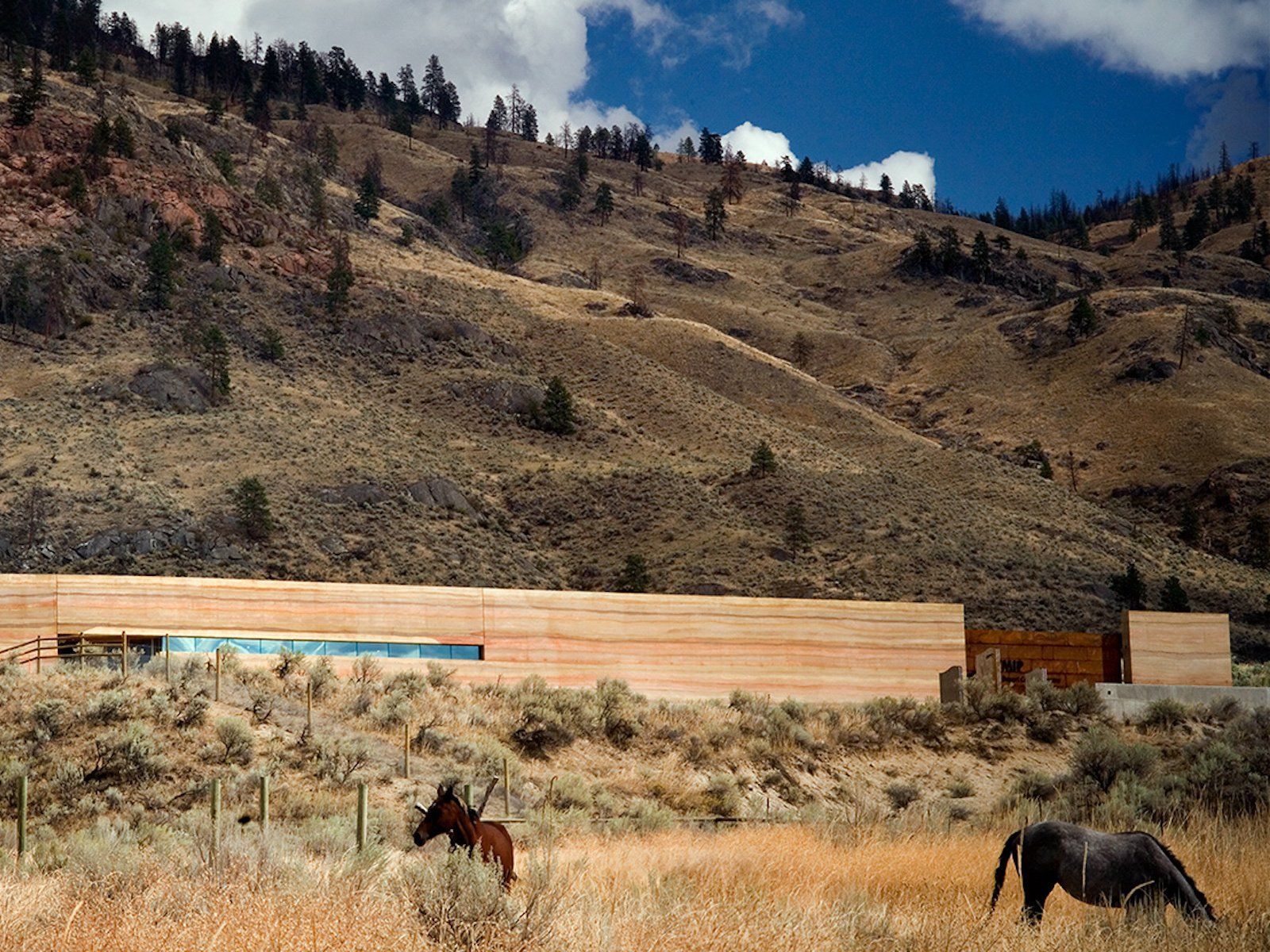 nk mip desert landscape with SIREWALL rammed earth building nestled between the ground and the mountain and blue sky, two horses are grazing in the front