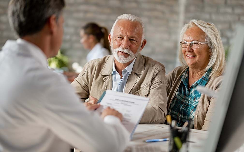 Doctor reviewing paperwork with senior couple at a desk; smiling, light-colored clothing.