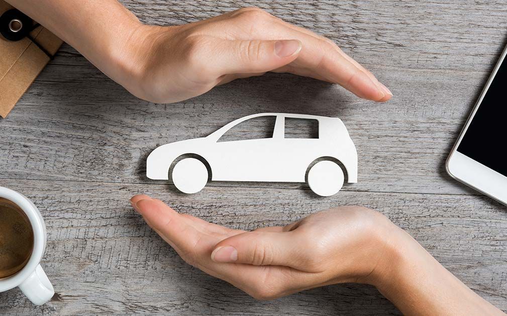 Hands cradling a white paper car on a wooden desk, suggesting protection or insurance.