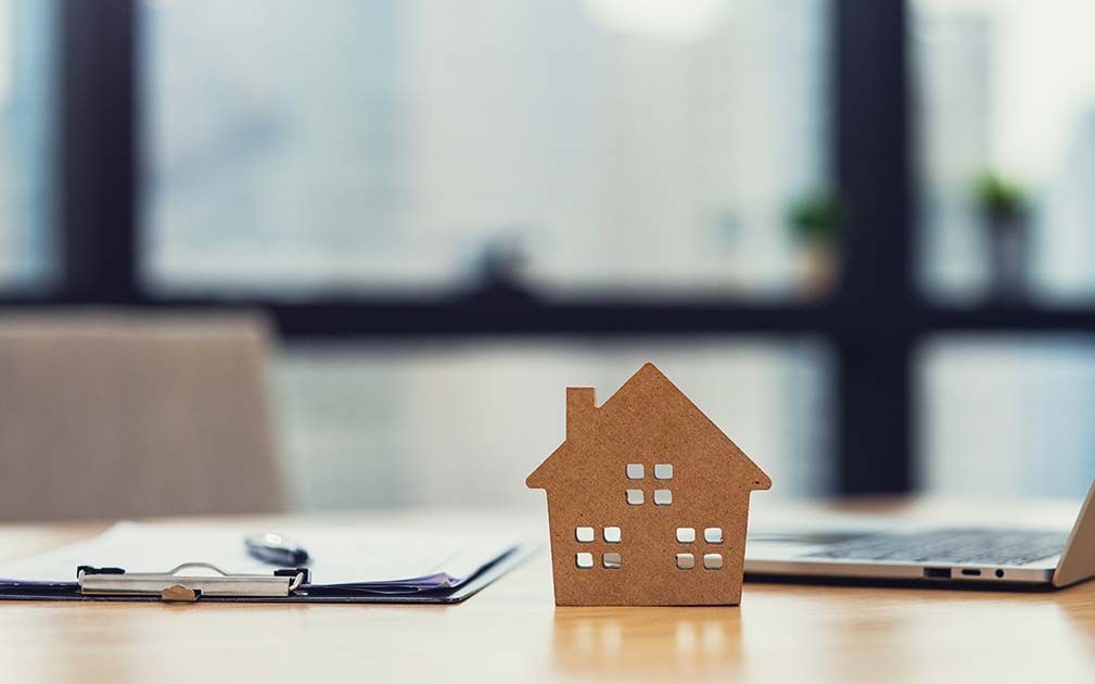 Wooden house model on desk with clipboard, pen, and laptop.