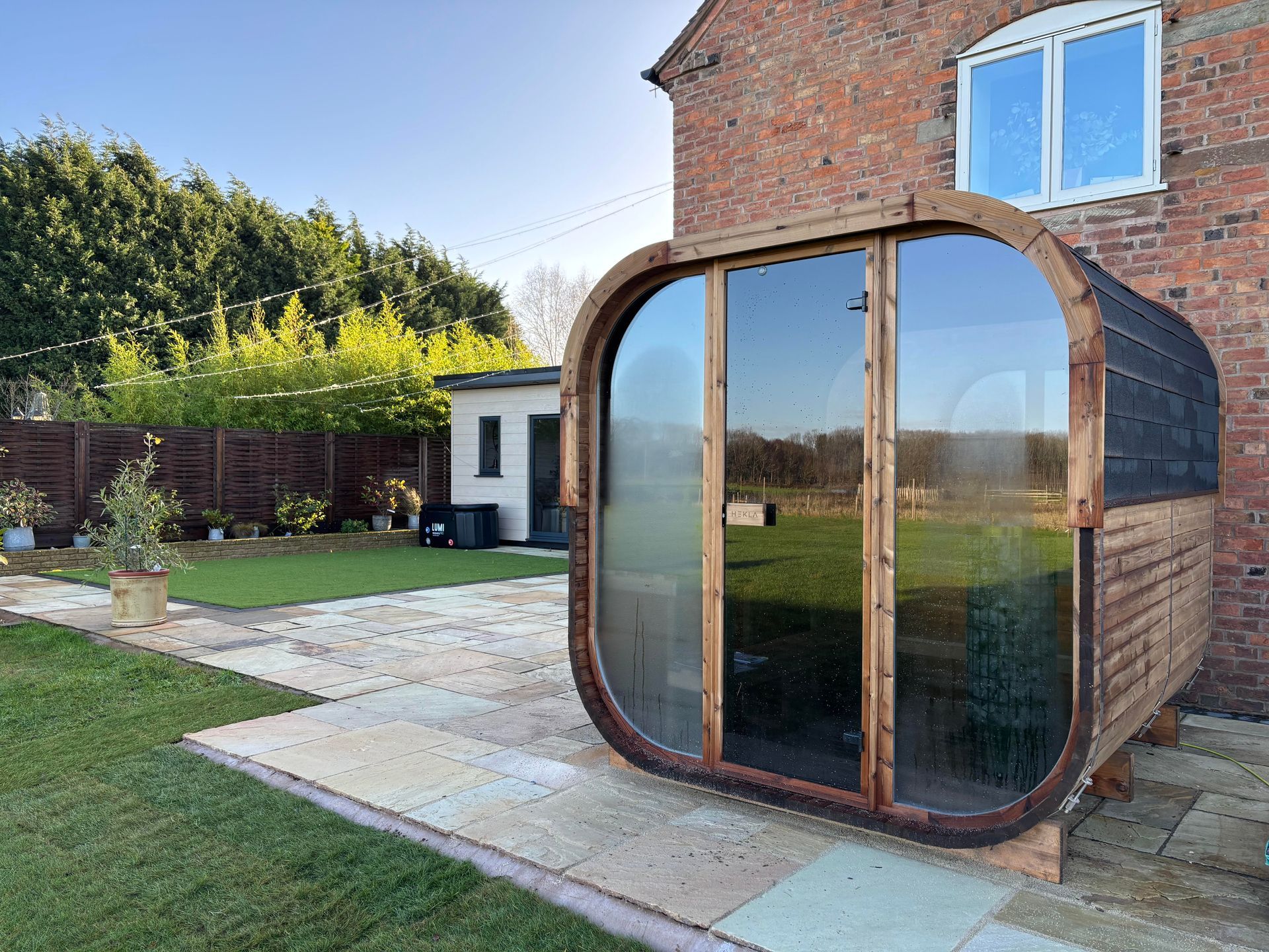 Backyard garden with a wooden sauna pod beside a brick house and lawn
