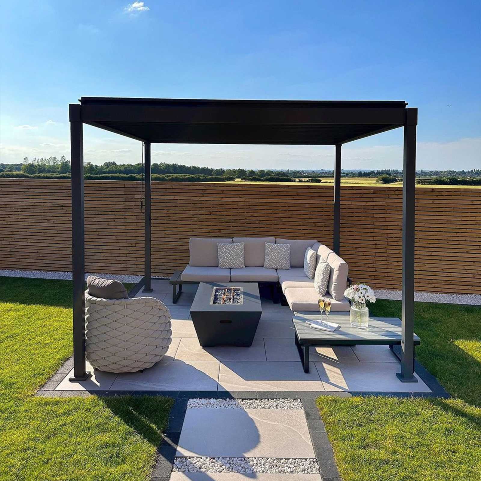 Modern outdoor lounge with pergola, beige seating, coffee table, and fire pit on a patio with grass and wood fence.