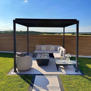 Modern outdoor lounge with pergola, beige seating, coffee table, and fire pit on a patio with grass and wood fence.