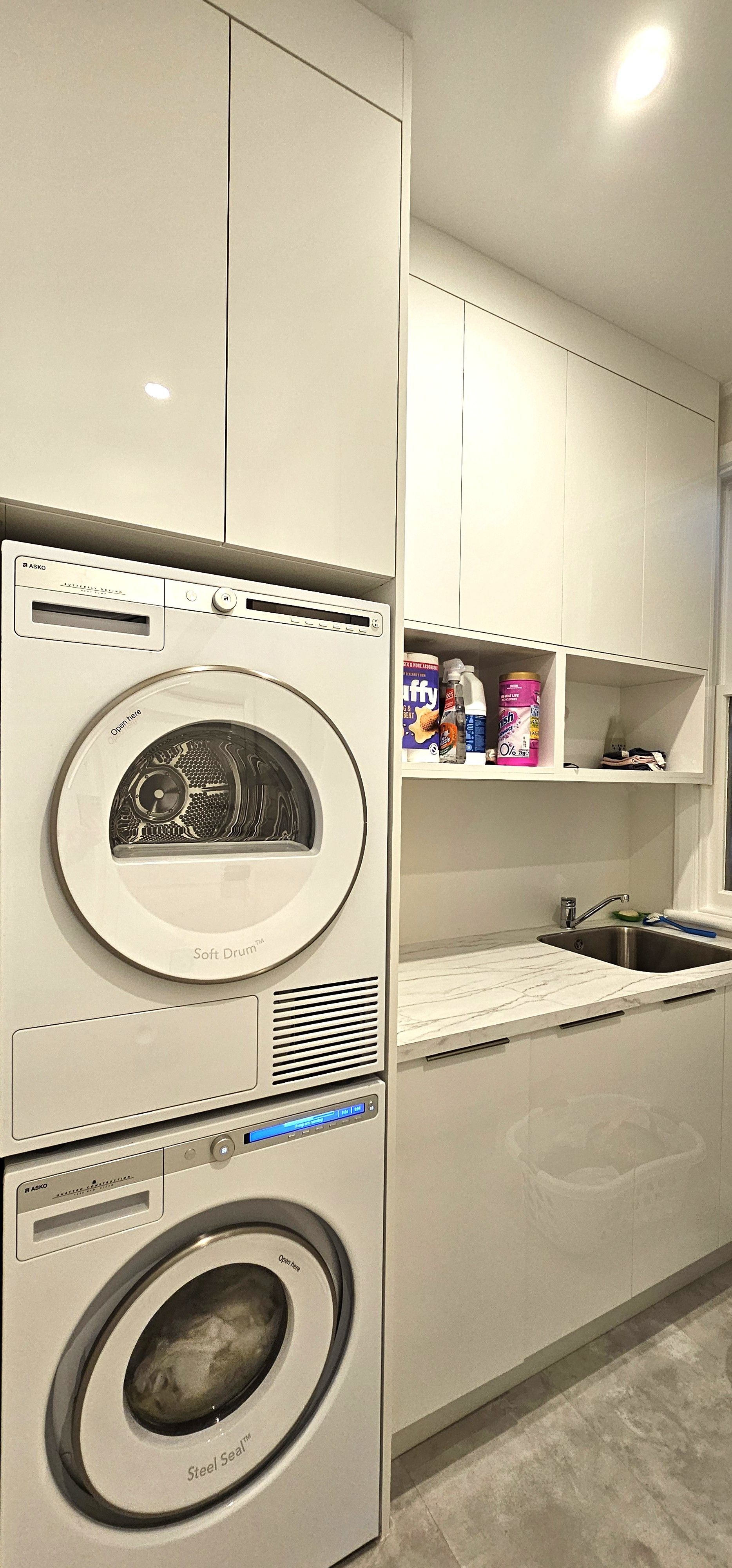 A white laundry room with stacked washer and dryer. Cabinets line the wall, along with a sink and countertop.