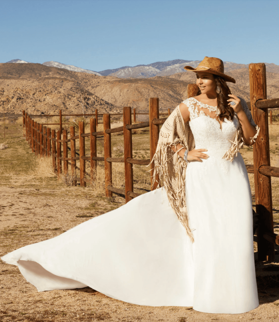 Eine Frau in einem weißen Kleid und einem Cowboyhut steht neben einem Holzzaun.