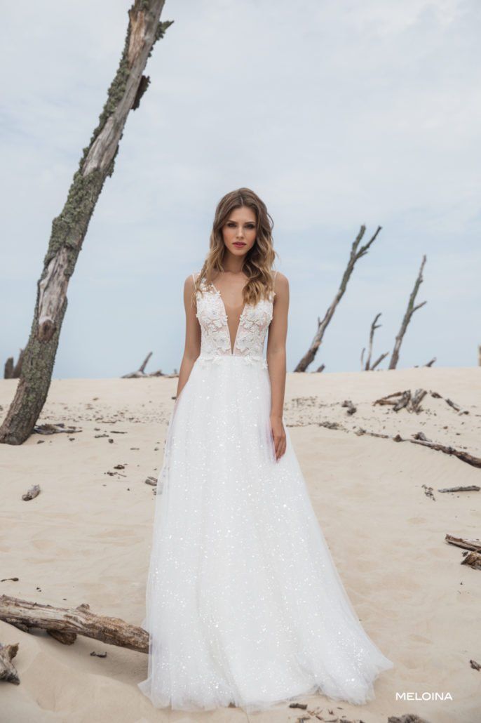 Eine Frau in einem weißen Hochzeitskleid steht neben einem Baum an einem Strand.
