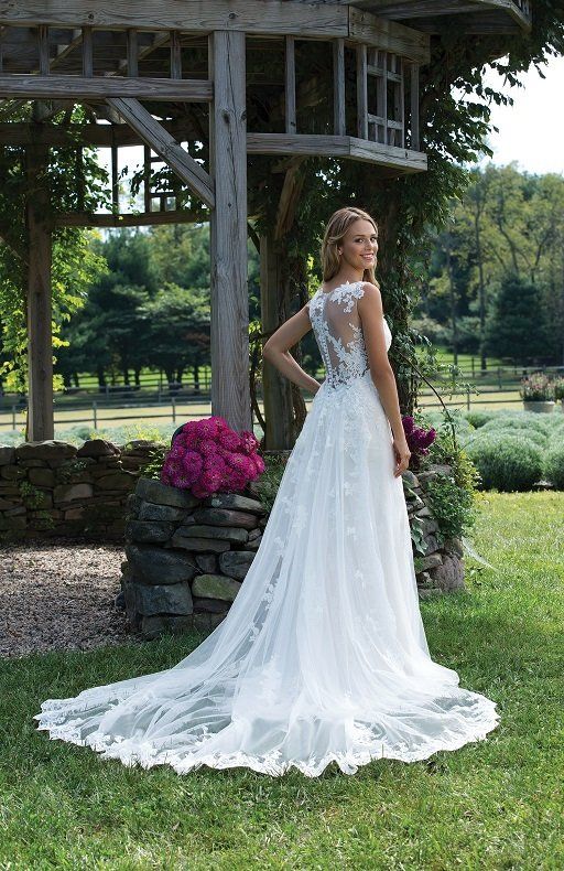 Eine Frau in einem weißen Brautkleid steht im Gras vor einer Holzkonstruktion.