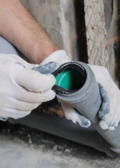 Hands in gloves fitting a rubber seal onto a gray plastic pipe, likely plumbing in a concrete setting.