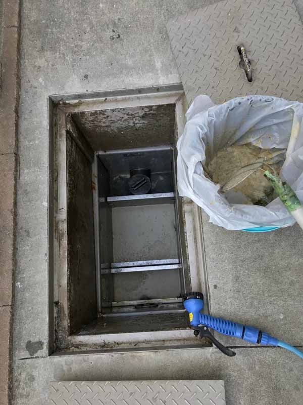 Open sewer access with a container of debris next to it and a blue hose. Concrete setting.