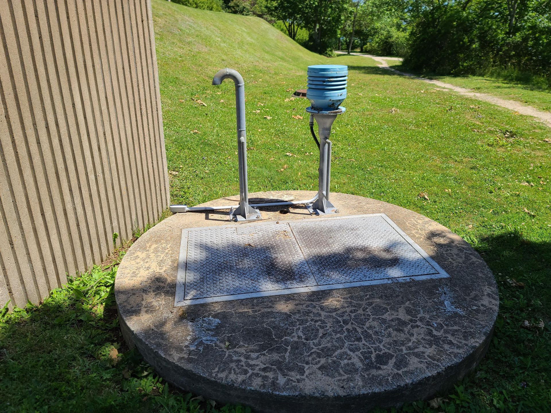 Concrete well cover with pump and faucet in grassy park next to a building.