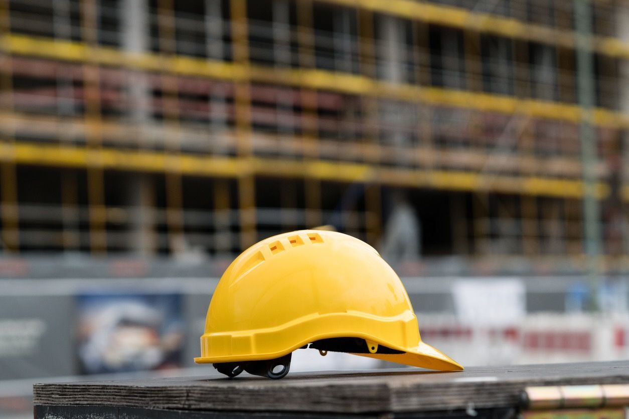 A yellow hard hat sits on a plank of wood. A construction site is visible in the background.