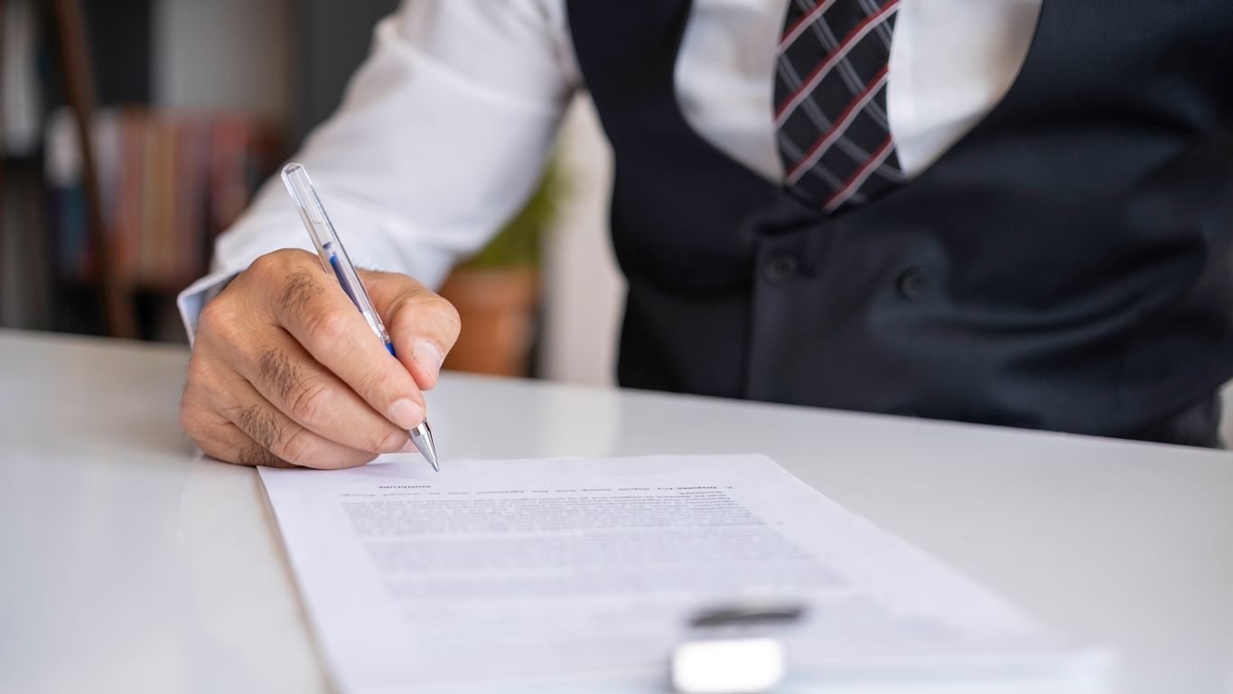 A man wearing business clothes signs a document with a pen.