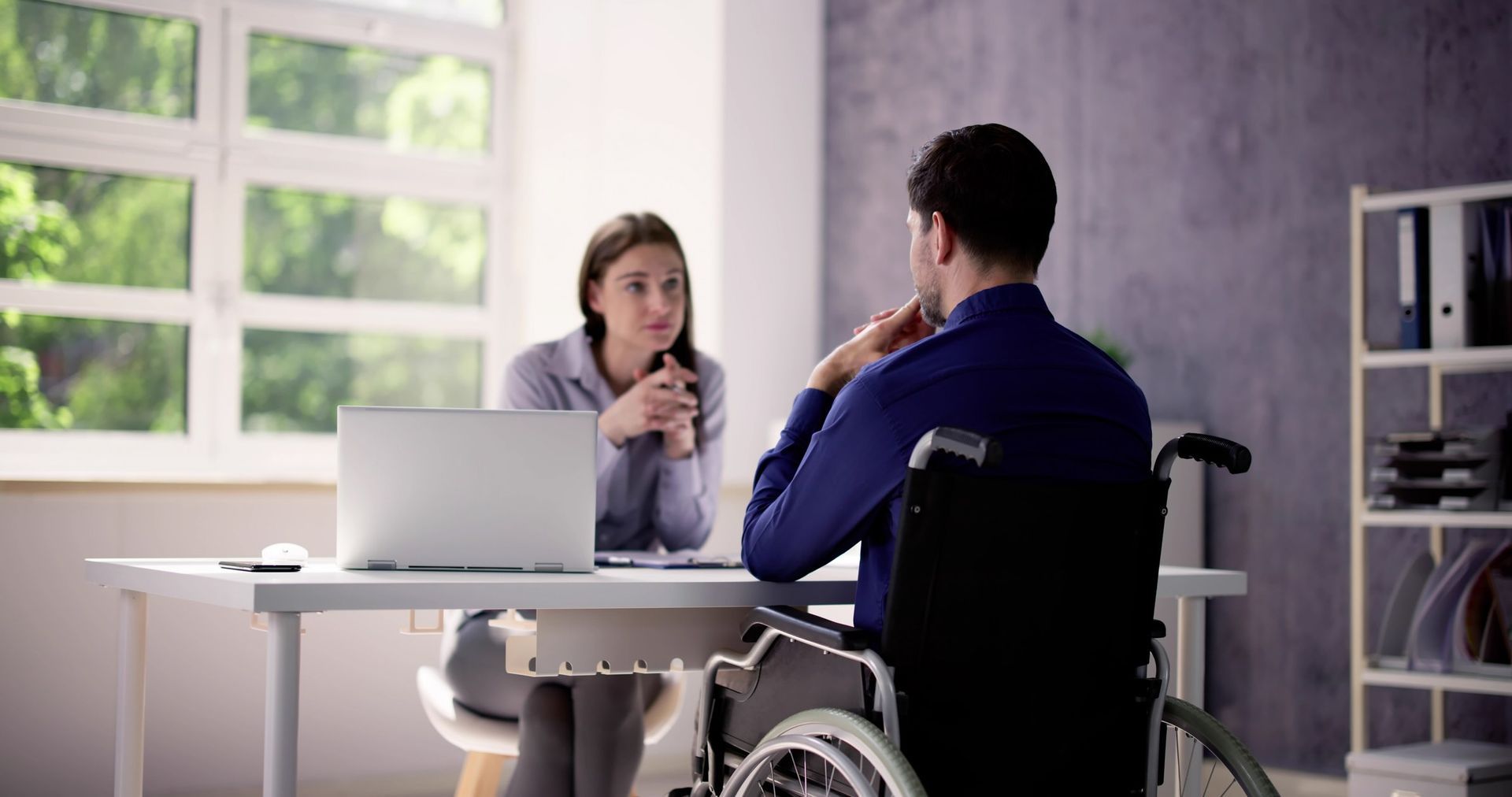 A woman sitting at a desk with a laptop open talks to a man using a wheelchair.