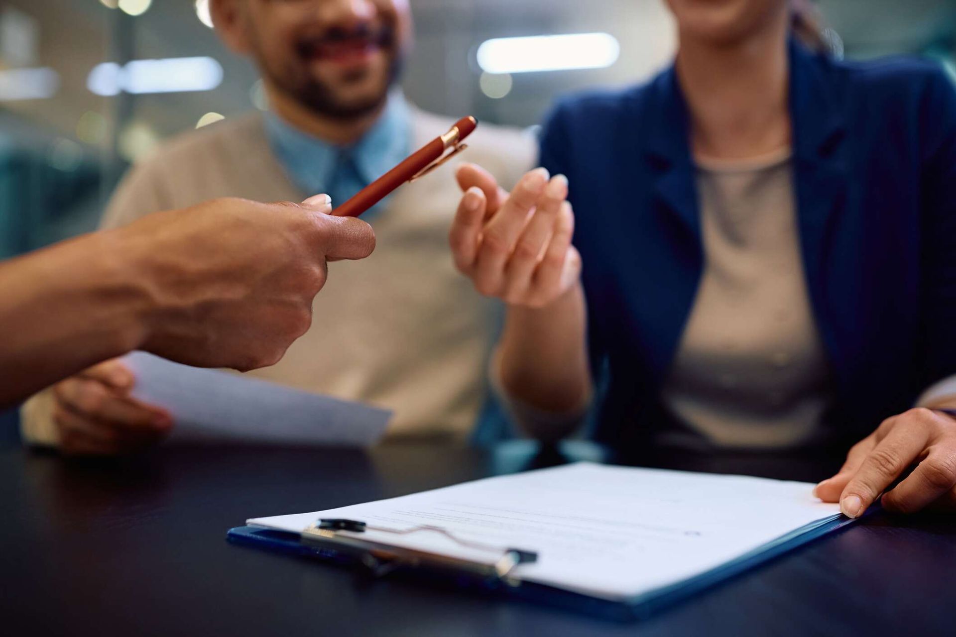 A man and a woman sit next to each other at a desk with a clipboard. Off-camera, a person hands the woman a pen.