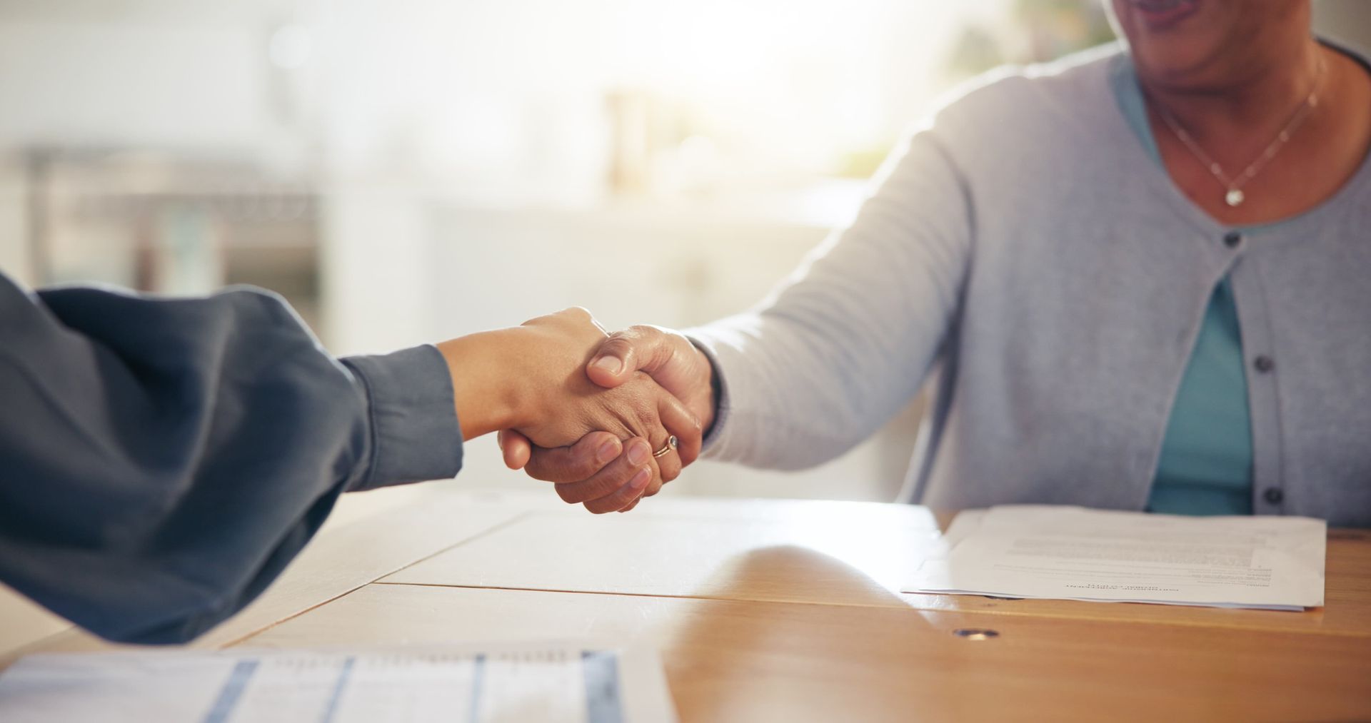 Two people shake hands while sitting at a table with printed documents.