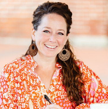 Woman in blazer leans on railing, smiling outdoors, green field in background.