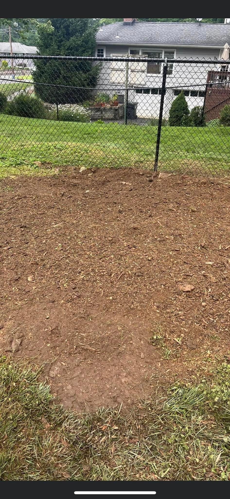 A cleared dirt patch in a grassy yard, with a fence and house in the background.