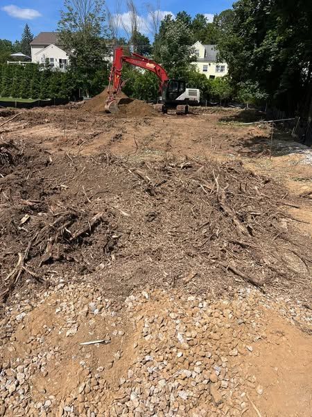 Construction site with excavator, cleared land, and debris. Houses in the background.
