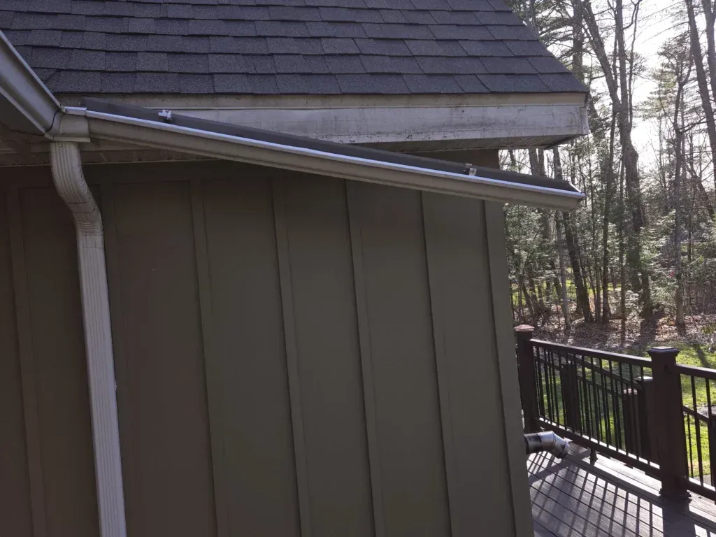 Green siding on a building with a dark roof, white gutter, and a deck with trees in the background.