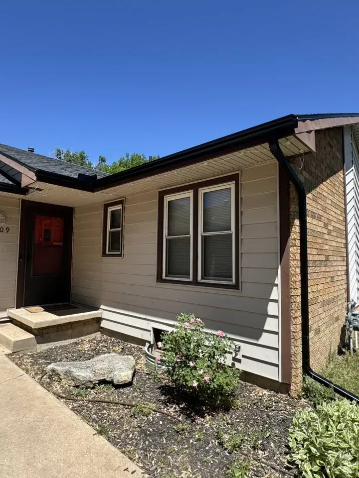 Tan house with brown trim, brick accent, windows, and a red door under a blue sky.