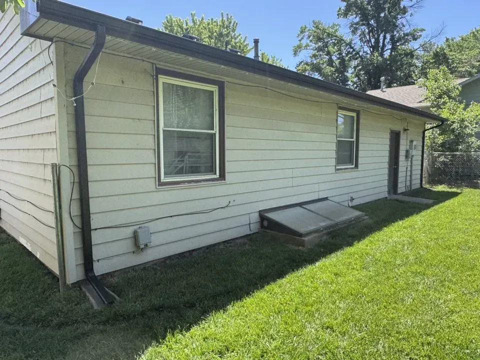 Side of a light-colored house with black gutters, windows, and a basement window well, in a grassy yard.