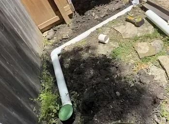 White PVC pipes laid in a trench along a wooden fence for an underground drain system, dirt and grass around.