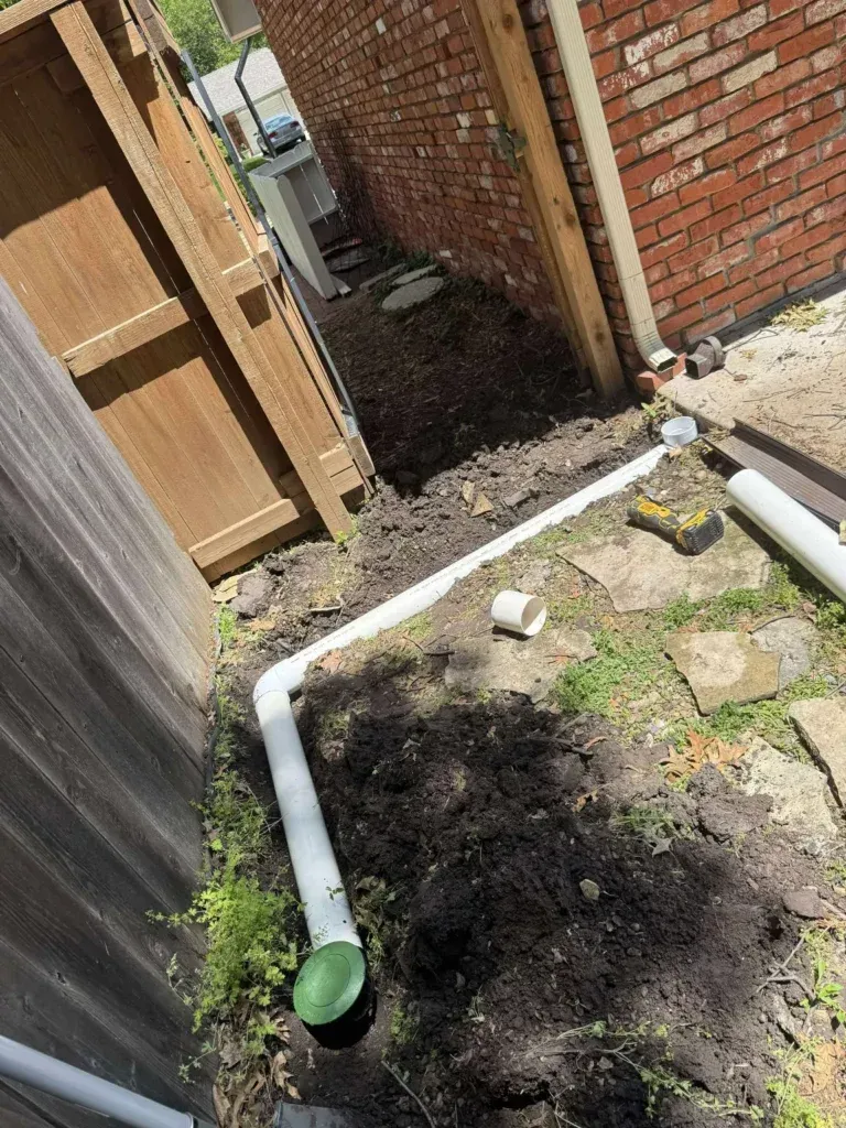 White PVC pipe snaking through dirt near a brick wall and wooden fence, for a drainage system.