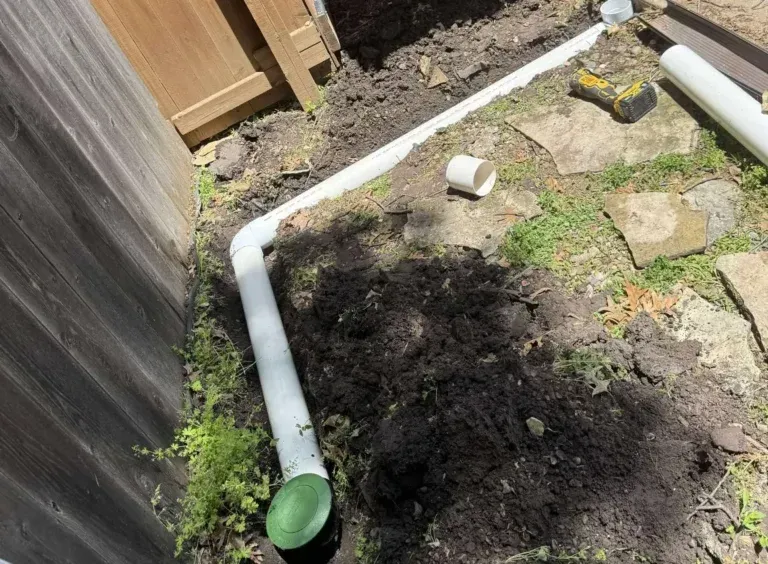 White PVC pipes laid in a trench along a wooden fence for an underground drain system, dirt and grass around.