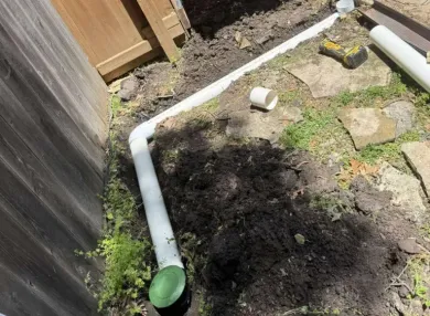 White PVC pipes laid in a trench along a wooden fence for an underground drain system, dirt and grass around.
