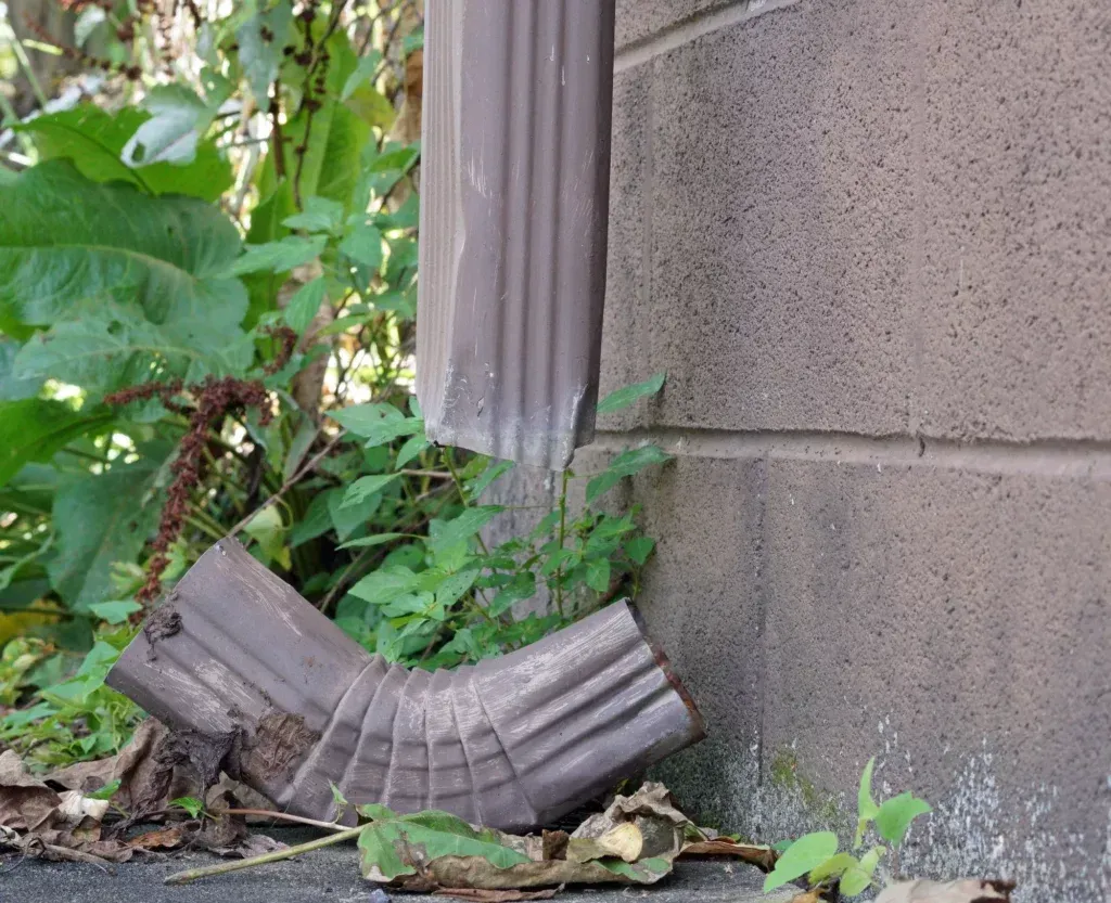 Brown gutter detached from a building, angled to the ground, with greenery around it.