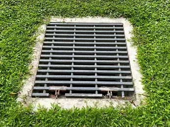 Metal grate over a catch basin drain in green grass, set in a concrete square.