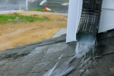 Rainwater pouring from a black gutter onto a gray shingled roof, outdoors.