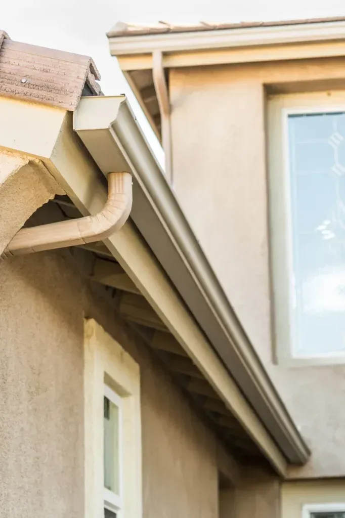 Beige gutters and downspout on a tan house with a brown roof, and a white window frame.