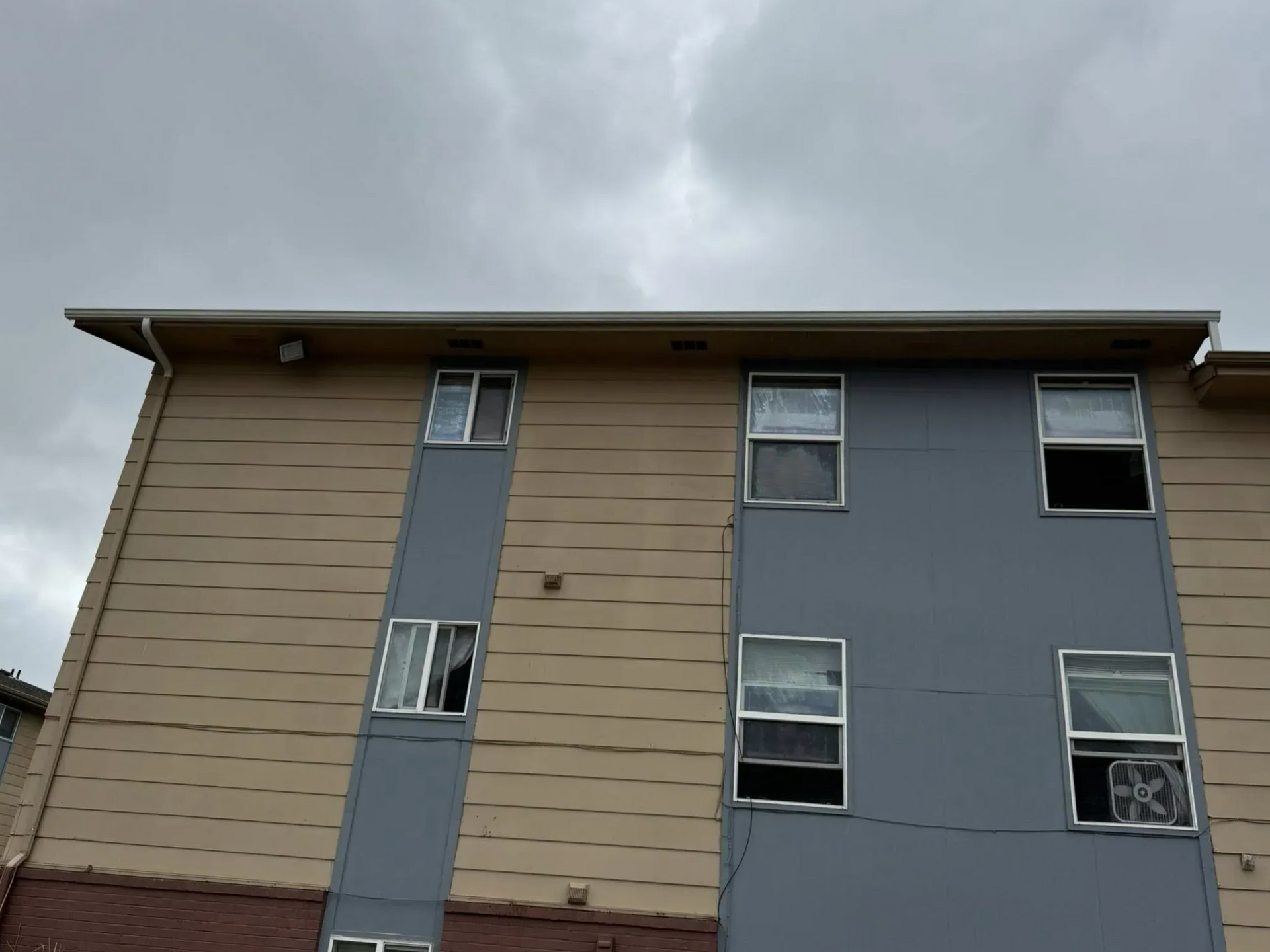 Exterior of a multi-story building with windows, gray and tan siding, against a cloudy sky.