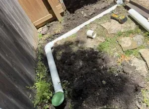Backyard corner with white PVC pipe underground drain ready to be buried, muddy ground, rocks, and a wooden fence