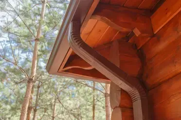 Brown seamless gutter and downspout matching a wooden house with trees in the background.
