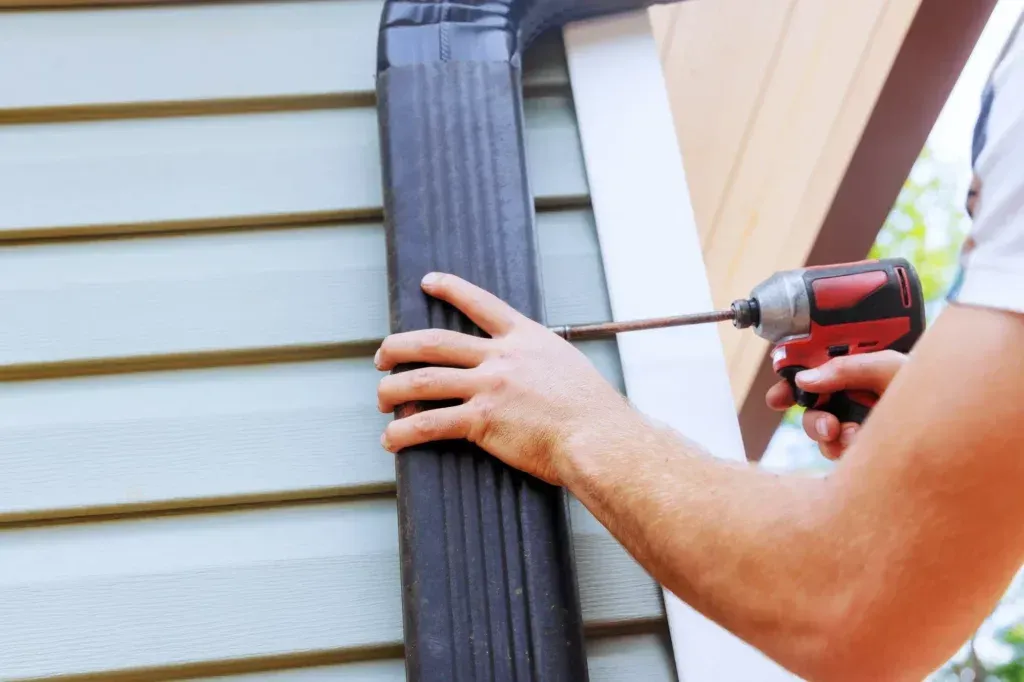 Person using a power drill to attach a black gutter downspout to a house with gray siding.