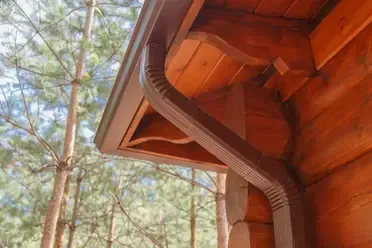 Brown gutter system on a wooden cabin, with pine trees in the background.
